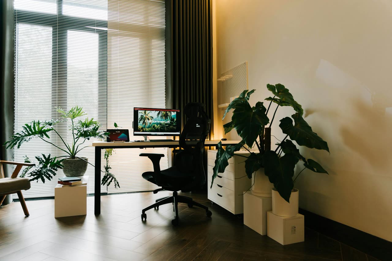 A small independent home office: a desk with a monitor, an ergonomic chair, and potted plants beside a large window.