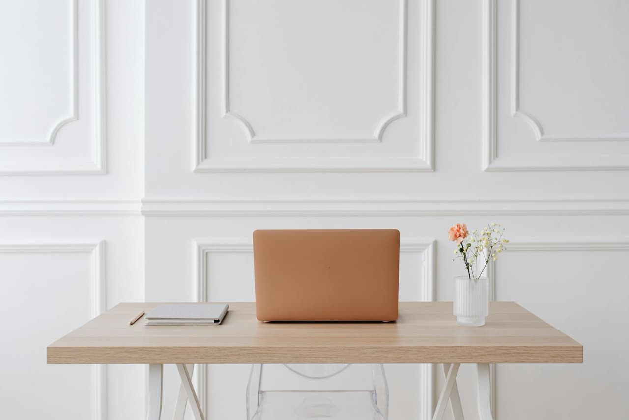 A laptop, a notebook, and a small vase of flowers on a minimal wooden desk against a white-panelled wall.